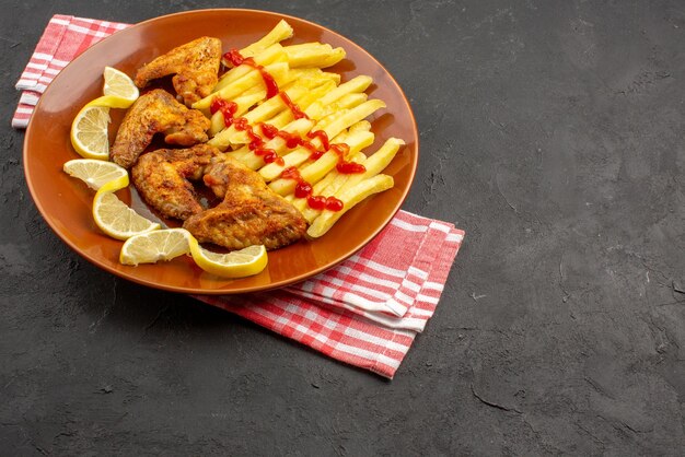 Side view chicken french fries on pink-white checkered tablecloth orange plate of appetizing french fries chicken wings ketchup and lemon on the left side of the dark table