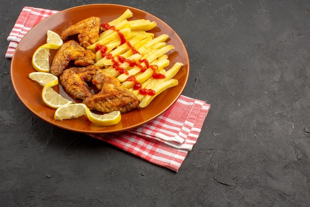 Side view chicken french fries on pink-white checkered tablecloth orange plate of appetizing french fries chicken wings ketchup and lemon on the left side of the dark table