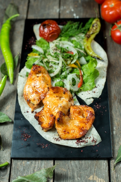 Side view of chicken kebab served with onions grilled tomato and pepper on black board