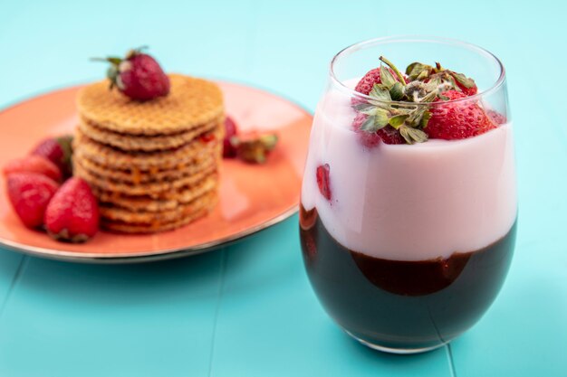 Side view of chocolate milkshake with strawberries with waffles on an orange plate on blue surface