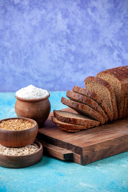 Side view of chopped in half black bread slices on wooden boards flour wheat oatmeal in bowls on light ice blue background