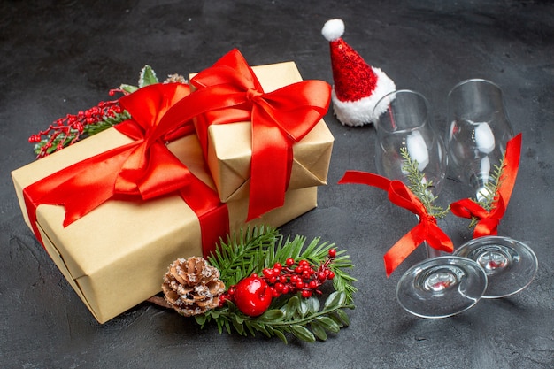 Side view of Christmas mood with beautiful gifts with bow-shaped ribbon and fir branches decoration accessories santa claus hat fallen glass goblets conifer cones on a dark background