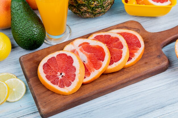 Вид сбоку цитрусовых как лимонный авокадо ананас с апельсиновым соком и нарезанный грейпфрут на разделочную доску на деревянном фоне