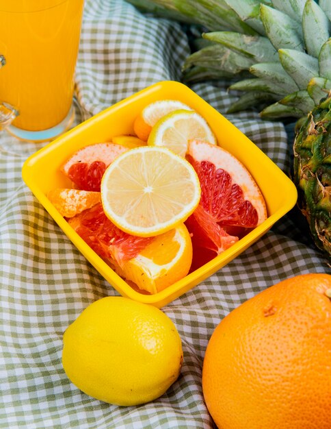 Side view of citrus fruits as lemon tangerine grapefruit kumquat in bowl with orange juice pineapple on plaid cloth background