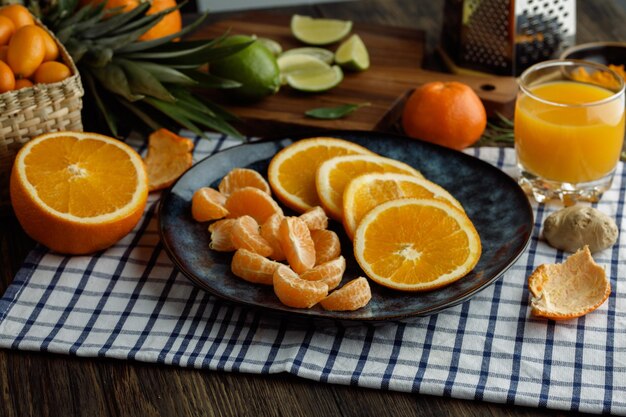 Side view of citrus fruits as orange and tangerine slices in plate with orange juice tangerine shell on plaid cloth with orange zest kumquats lime on wooden background