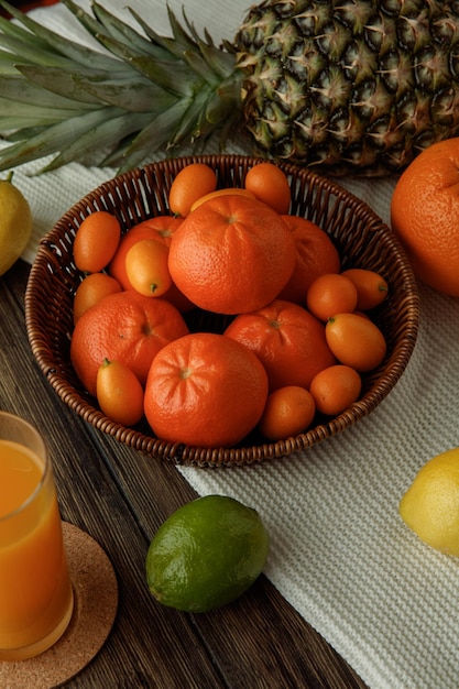 Side view of citrus fruits as tangerines and kumquats in basket lemon lime orange pineapple on cloth with orange juice on wooden background