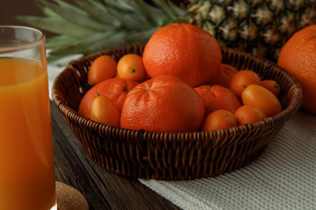 Вид сбоку на цитрусовые в виде мандаринов и кумкватов в корзине с апельсиновым ананасом на ткани с апельсиновым соком на деревянном фоне
