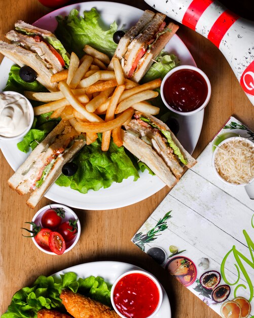 Side view of club sandwich with chicken french fries and sauces on wooden board