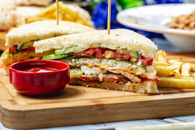 Side view club sandwich with grilled chicken tomato cucumber lettuce mayo ketchup and french fries on a board