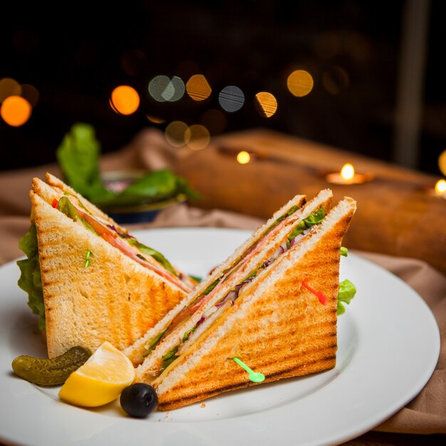 Side view club sandwich with salted cucumbers and lemon and olives in round white plate