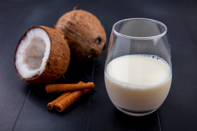 Side view of coconuts with a glass of milk and cinnamon sticks on black surface