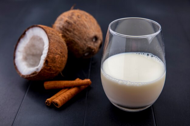 Вид сбоку кокосовых орехов со стаканом молока и палочки корицы на черной поверхности