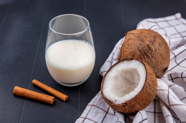 Side view of coconuts with a glass of milk with cinnamon sticks on a checked tablecloth on a wooden surface