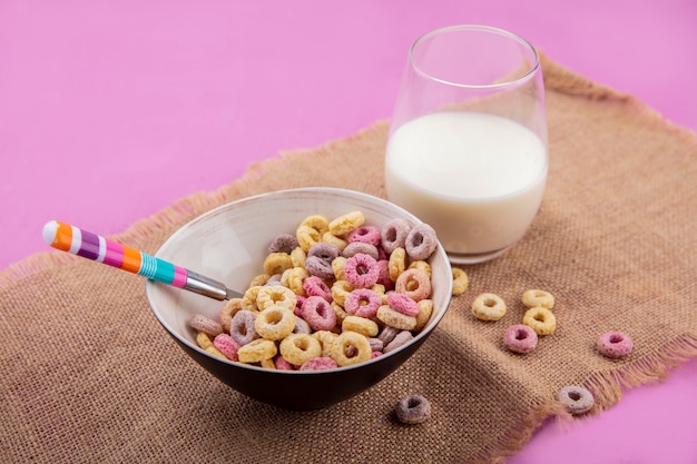 Vista laterale dei cereali colorati su una ciotola con un cucchiaio con un bicchiere di latte sul panno di sacco sulla superficie rosa