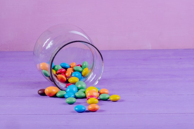 Free photo side view of colorful candies scattered from a glass jar on purple background with copy space