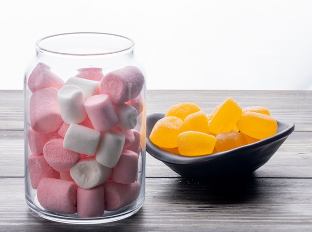 Side view of colorful marshmallow in a glass jar and marmalade candies in a bowl on wooden table