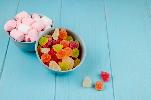 Side view of colorful tasty marmalade candies and marshmallows in bowls on blue