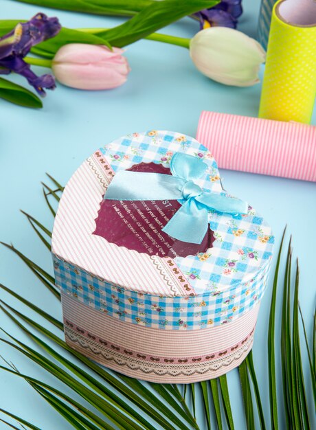 Side view of colorful tulip flowers and heart shaped gift box with rolls of adhesive tape on blue background