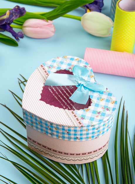 Side view of colorful tulip flowers and heart shaped gift box with rolls of adhesive tape on blue background