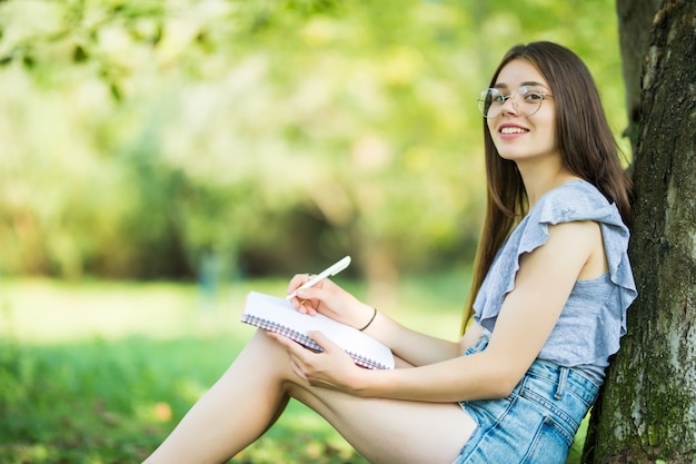 Side view of concentrated brunette woman in eyeglasses sitting near the tree in park and writing something on notebook