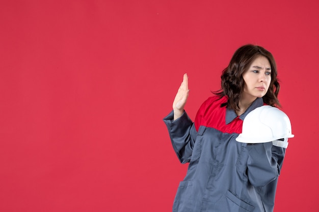 Side view of concerned female architect holding hard hat and standing on isolated red background