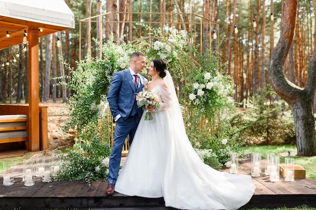 Side view of confident groom dressed in elegant suit keeping hand in pocket and looking at bride which standing opposite him