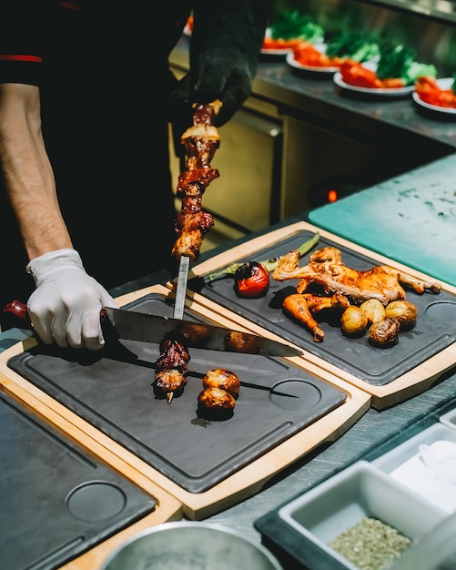 Side view the cook removes from a skewer tike kebab on a board with grilled chicken and vegetables