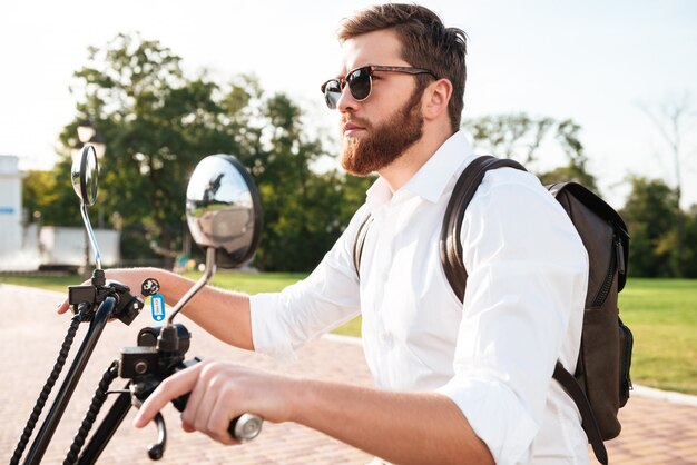 Side view of Cool bearded man in sunglasses with backpack rides on modern motorbike outdoors and looking away