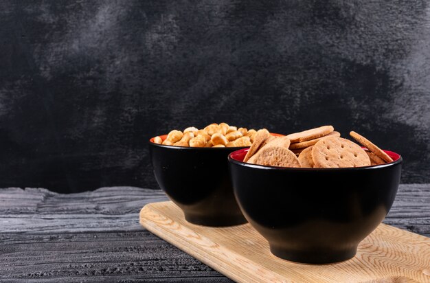 Side view of crackers in bowls on wooden cutting board and dark  horizontal