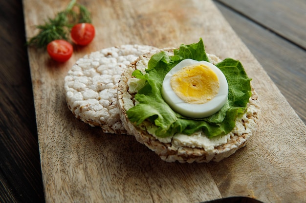Side view of crispbreads with lettuce and egg slice on one of them and half cut tomato with dill on cutting board on wooden background