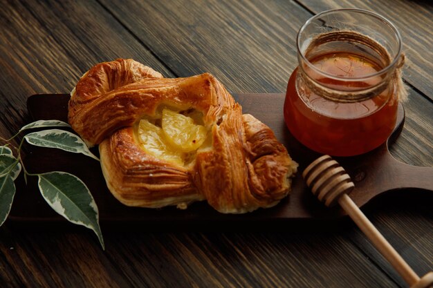 Side view of croissant and glass jar of jam with leaves on cutting board on wooden background