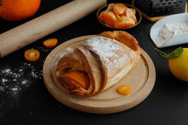 Vista laterale del croissant su sottopentola con farina di kumquat limone fette di mandarino e mattarello su sfondo nero