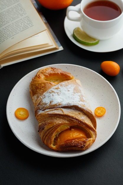 Side view of croissant with kumquat slices in plate and cup of tea open book with whole kumquats on black background