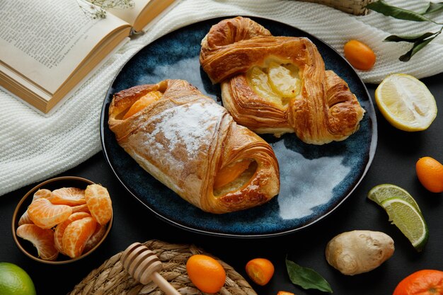 Side view of croissants in plate with tangerine slices and open book with basket of kumquat and little flower on book with ginger lime slices trivet and honey dipper on cloth on black background