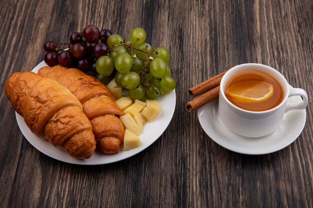 Side view of croissants with grapes and cheese slices in plate with cup of hot toddy with cinnamon on saucer on wooden background