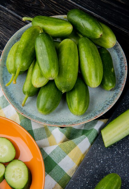 Side view of cucumbers in plate with cut one on cutting board and sliced one in plate on cloth and wooden table