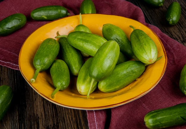 Side view of cucumbers in plate with other ones on bordo cloth and wooden table