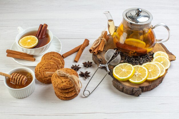 Side view of a cup of black tea cookies honey and herbal tea in a glass pot and chopped lemon cinnamon limes on a wooden tray on white background