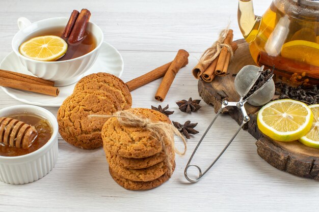 Side view of a cup of black tea stacked cookies honey and herbal tea in a glass pot and chopped lemon cinnamon limes on a wooden tray on white background