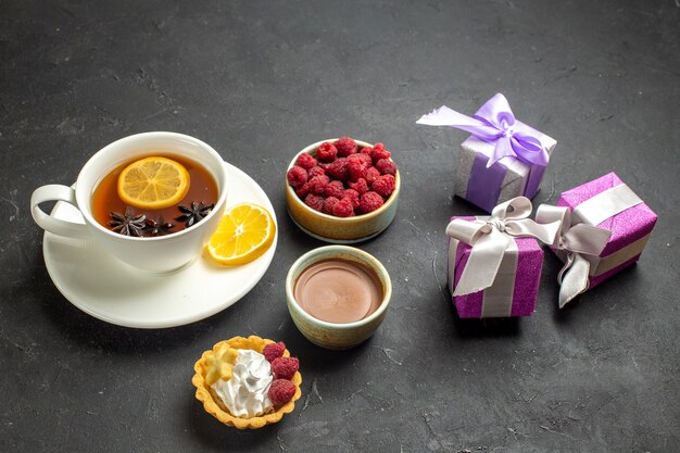 Side view of a cup of black tea with lemon served with chocolate raspberry and gifts on dark background