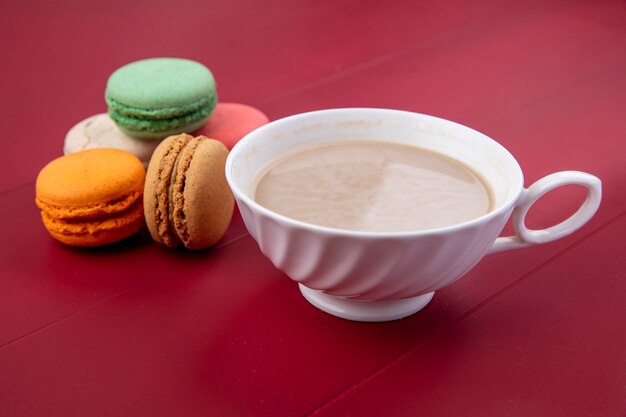 Side view of cup of cappuccino with colored macarons on a red surface