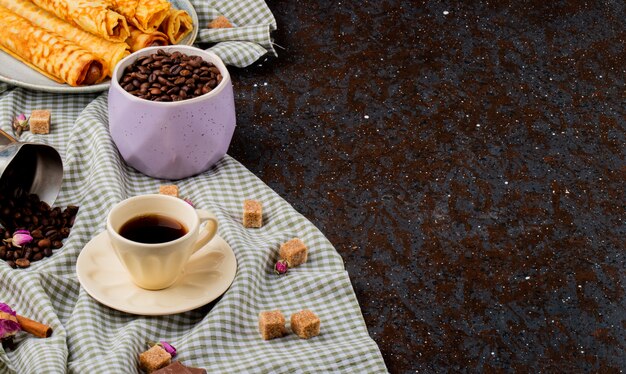 Side view of a cup of coffee and brown sugar cubes chocolate and coffee beans scattered on the plaid tablecloth with copy space