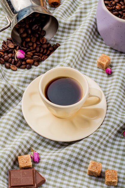 Вид сбоку чашки кофе и коричневого сахара кубиками шоколада и кофейных зерен, разбросанных на клетчатой скатерти