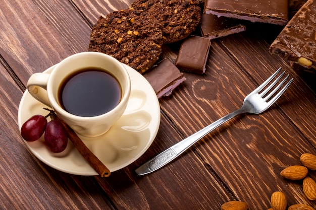 Side view of a cup of coffee with chocolate bar and oatmeal cookies with fork on wooden background