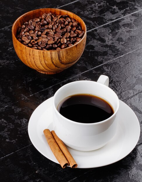 Side view of a cup of coffee with cinnamon sticks and a wooden bowl with coffee beans