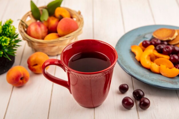 Side view of cup of coffee with plate of pancakes and apricot slices with cherries and basket of apricot on wooden background