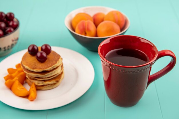 Side view of cup of coffee with plate of pancakes and apricot slices with cherries and bowls of cherry and apricot on blue background