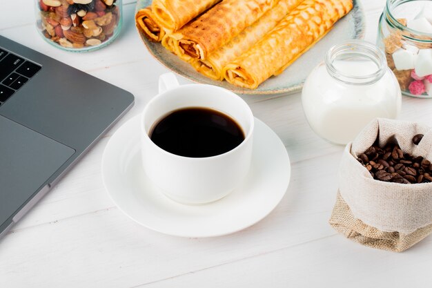 Side view of a cup of coffee with wafer rolls and laptop on white background
