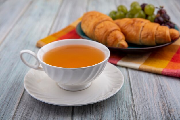 Side view of cup of hot toddy on saucer with croissants and grapes in plate on plaid cloth on wooden background