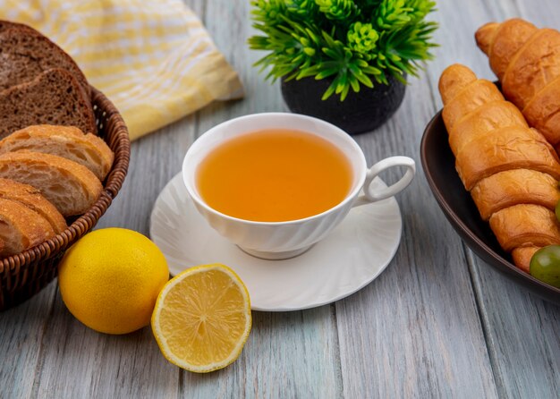 Side view of cup of hot toddy on saucer with rye crusty bread slices in basket and croissants in bowl with half cut lemon and plant on wooden background